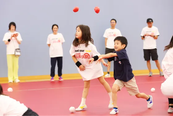 子どもたちがボールを投げながら楽しそうに遊んでいる。周りでは大人たちが微笑ましく見守っている。