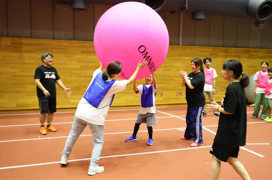 女の子と母親が協力しながら、大きなピンク色のゴムボールを持ち上げて一生懸命に運んでいる。