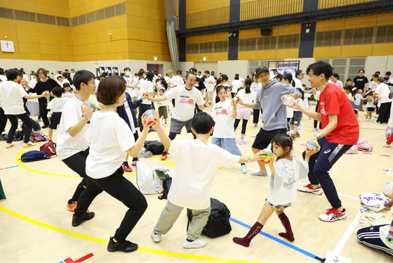 子どもと大人たちが楽しそうにエクササイズしている様子。皆んなで大きな輪をつくりながらバランスをとっている。