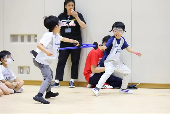 ゴーグルをつけた二人の男の子が向かい合い、スポンジの剣を使って真剣な眼差しでフェンシングの体験をしている。